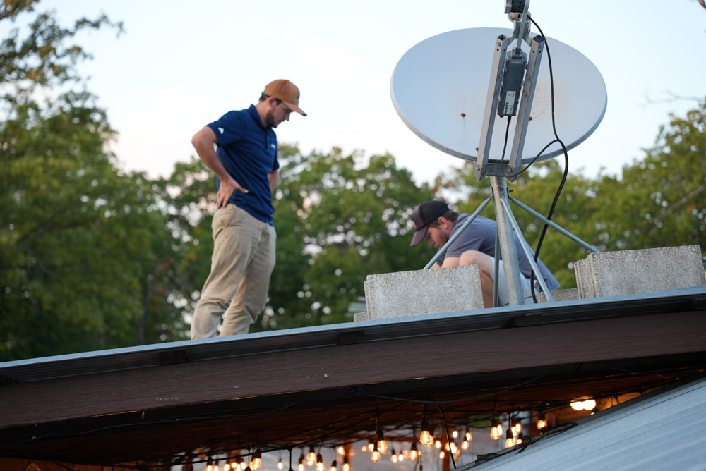 Two technicians are on a flat commercial roof during the day. One man stands while the other crouches to work near the base of a large satellite dish, preparing for an installation. String lights are visible hanging below the roof's edge.