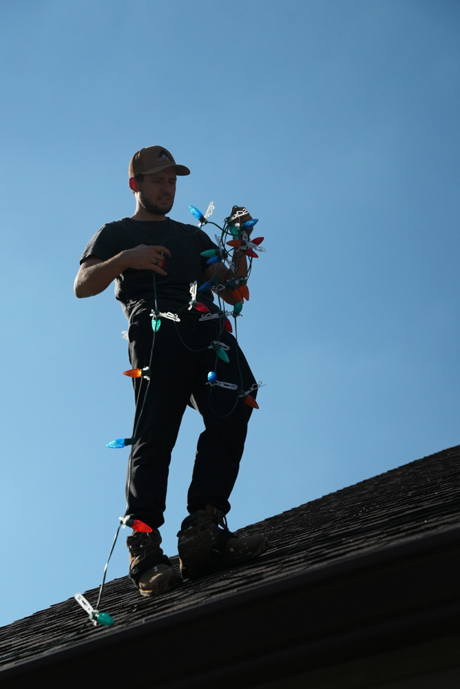 A full-body shot of a worker standing on a shingle roof, silhouetted against the bright sky. He is holding and untangling a large bundle of multi-colored Christmas lights, getting them ready for installation.