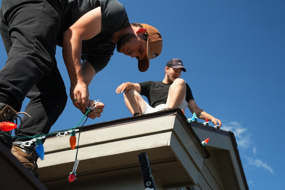 Two technicians collaborate on a rooftop under a clear blue sky. One man leans over the edge, securing a string of large, colorful bulbs, while his partner sits nearby, assisting with the professional holiday light installation.