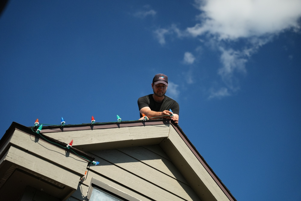 A low-angle shot looking up at a technician on the corner of a roof against a vibrant blue sky. He is carefully attaching a string of multi-colored Christmas lights to the edge of the roofline, preparing for a festive installation.